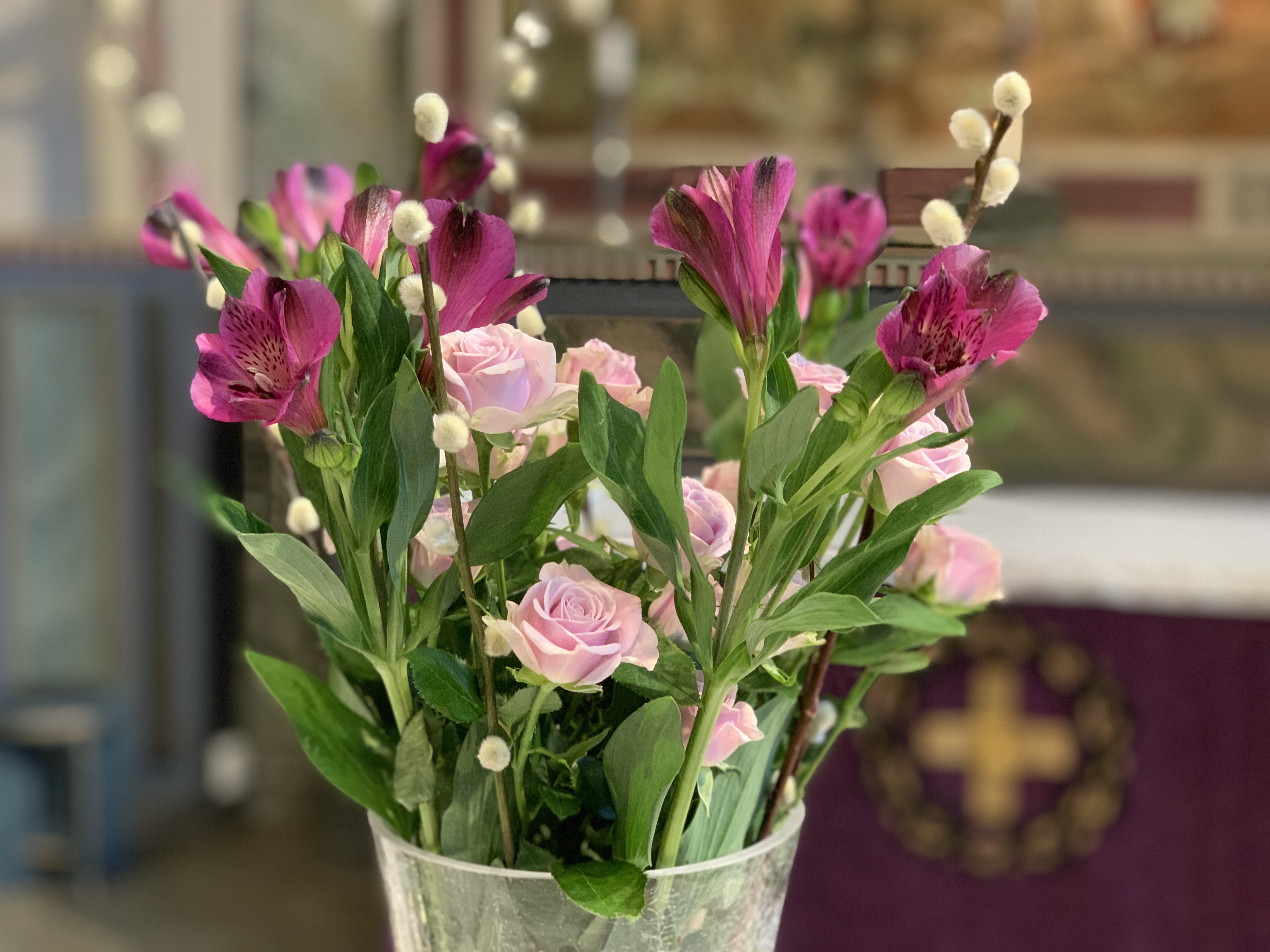 Lila och rosa altarblommor i en vas. I bakgrunden fastans lila altarduk med ett gyllene kors.