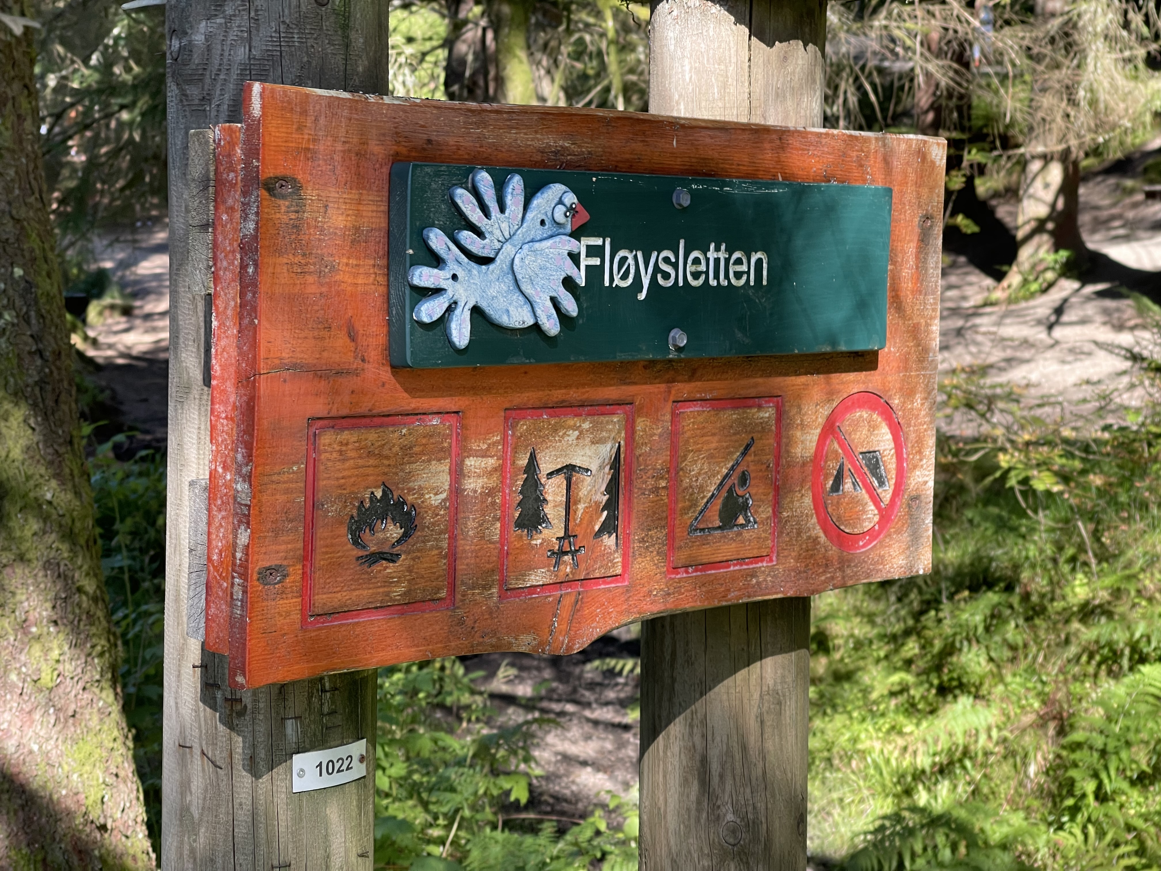 En skylt fäst vid två tjocka trästolpar mitt i skogen. Överst på skylten syns en vit duva och platsnamnet Fløysletten. I den nedre delen fyra ikoner: lägereldplats, pausbord, skyddstak och ingen camping.